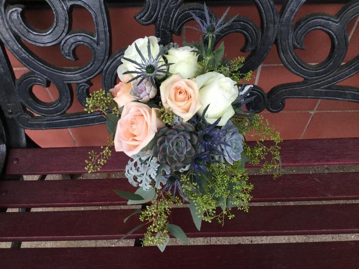 This bouquet designed with light peach and ivory roses, succulents, seeded eucalyptus, dusty miller and light blue thistle.