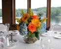 Blue hydrangea, orange yellow gerbera daisies, orange roses, solidago arranged in a rose bowl with river rock.