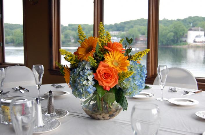 Blue hydrangea, orange yellow gerbera daisies, orange roses, solidago arranged in a rose bowl with river rock.